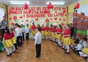 Chłopiec w białej koszuli i czarnych spodniach stoi na środku sali z pałeczką dyrygenta w ręku, w półkolu grają na instrumentach dziewczynki w czerwonych podkoszulkach i żółtych spódniczakach oraz chłopcy w białych koszulach i czarnych spodniach